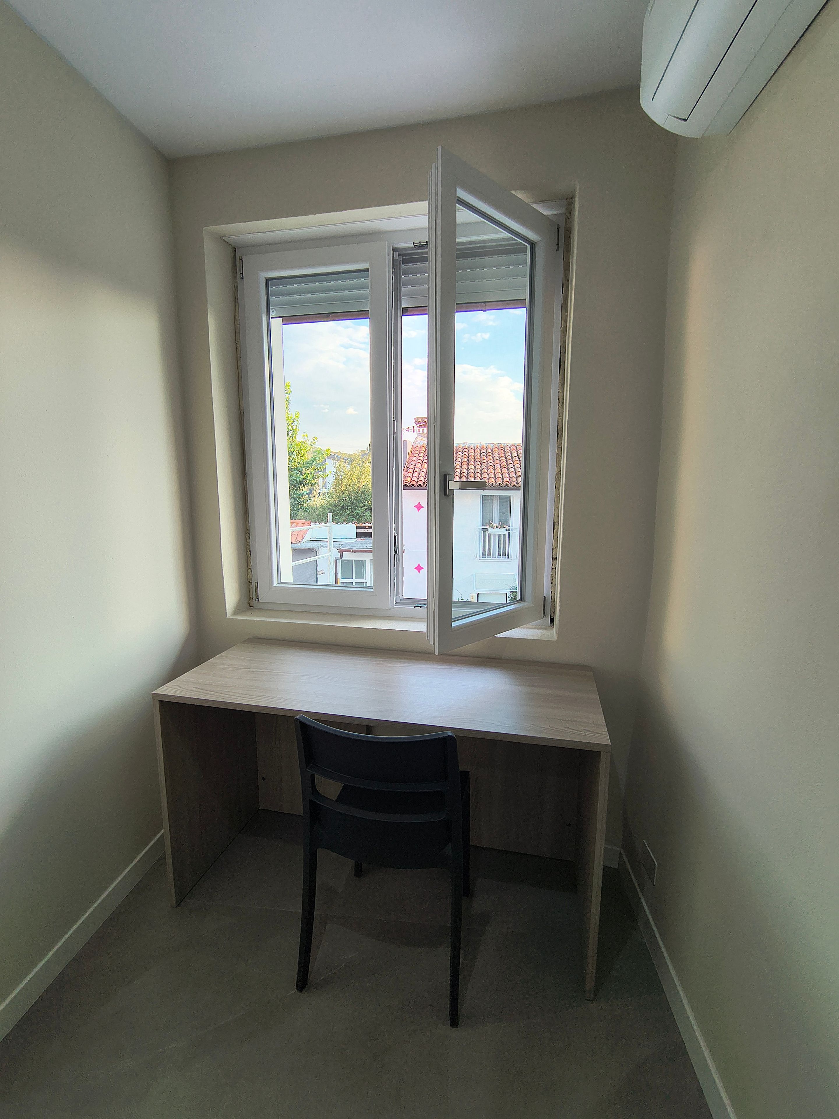 Ein Eckzimmer mit Schreibtisch und Stuhl, ein Fenster mit Blick auf Gebäude und einer Decken-Klimaanlage.