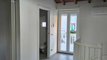 Une pièce avec un radiateur blanc, une porte blanche donnant sur une salle de bain, un escalier blanc et une table sur laquelle se trouve une paire de ciseaux.