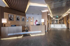 A hotel reception area with a wooden reception desk, a receptionist, and a wall with clocks and framed pictures. - Grand Plaza Tabuk (Tabuk)