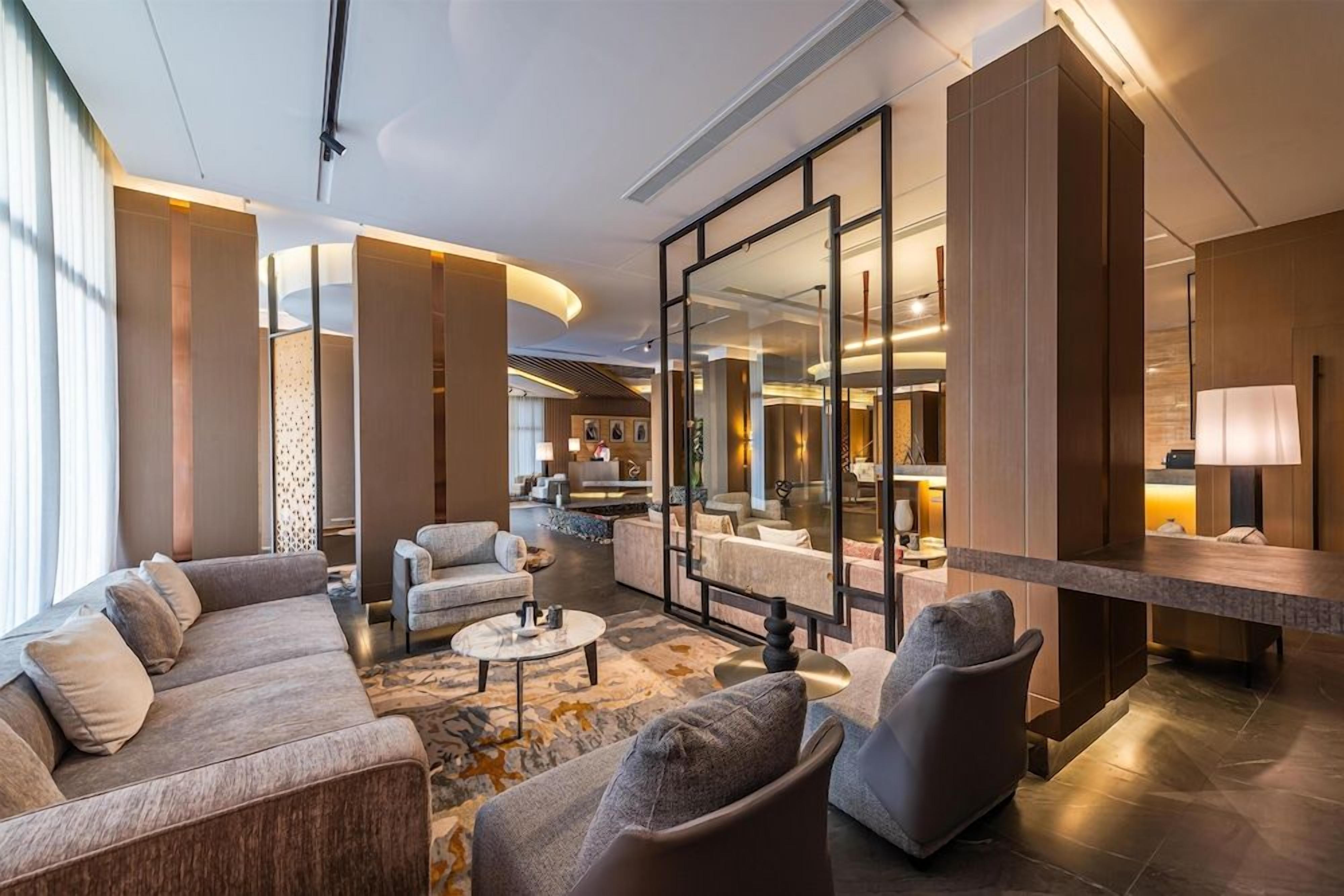 Um hall de hotel moderno com área de estar, balcão de recepção e um espelho grande refletindo o interior.