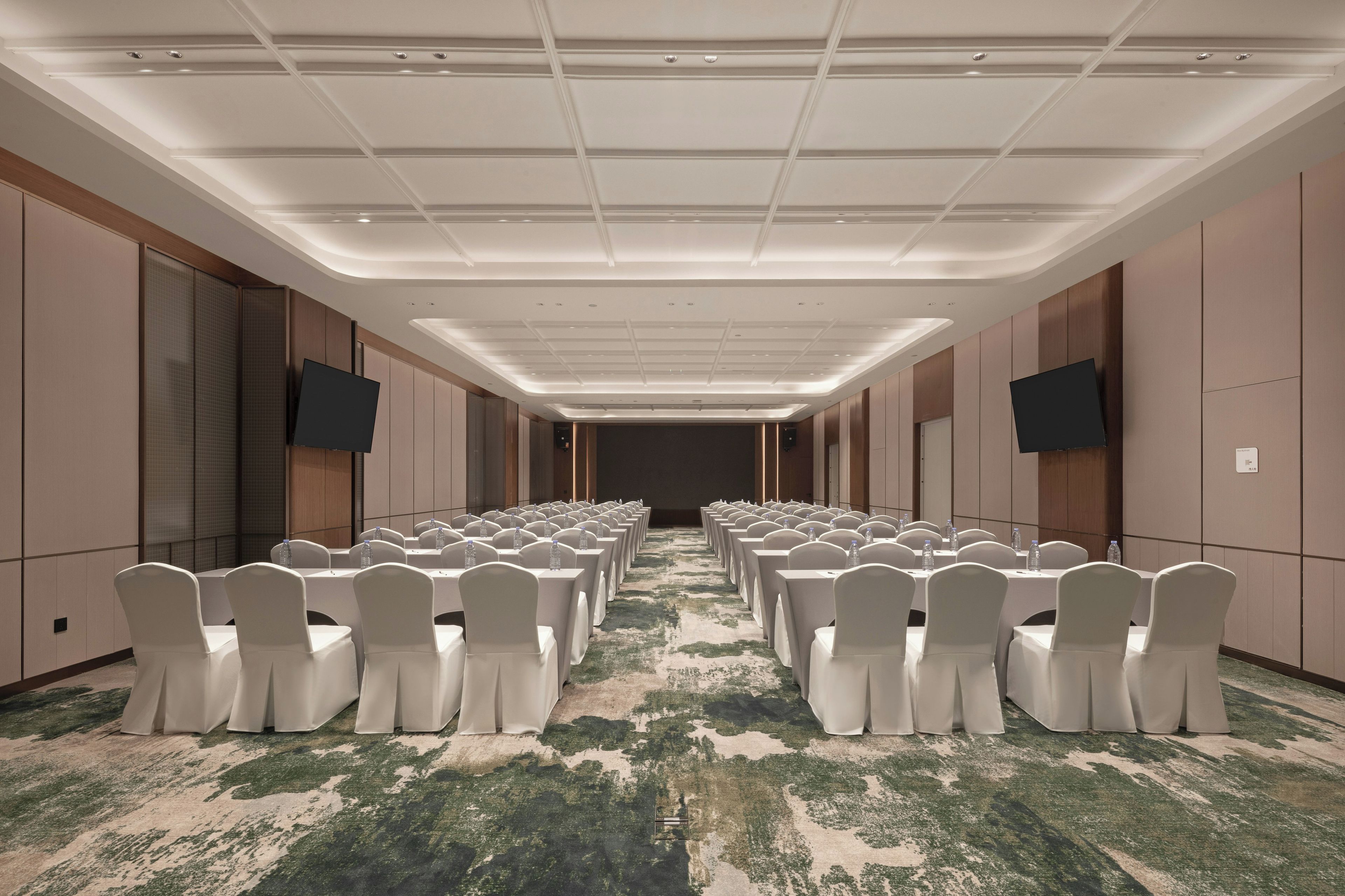 Un salón de conferencias con filas de mesas y sillas, equipado con botellas de agua y piso alfombrado.