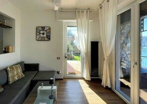 Sala de estar com sofá, mesa de centro e relógio na parede. Há uma porta de vidro deslizante que dá para uma área externa.