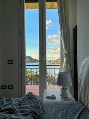 Ein Schlafzimmer mit einem Bett, einem Nachttisch mit Lampe und Blick auf eine Küstenlandschaft durch eine geöffnete Tür.