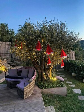 Uma árvore com decorações e luzes vermelhas, uma cadeira de vime e um deck de madeira.