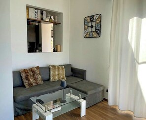 Ein modernes Wohnzimmer mit einem grauen Sofa, einem Glastisch, einer Wanduhr und einem Spiegel über einem Regal mit verschiedenen Gegenständen.