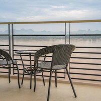 A balcony with wicker chairs and tables, overlooking a calm body of water and distant mountains.