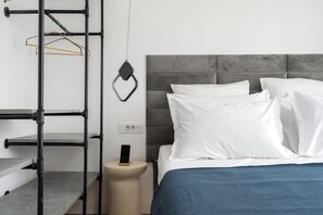 Une chambre moderne avec une tête de lit grise, du linge de lit blanc et une étagère en métal où sont rangés des vêtements.