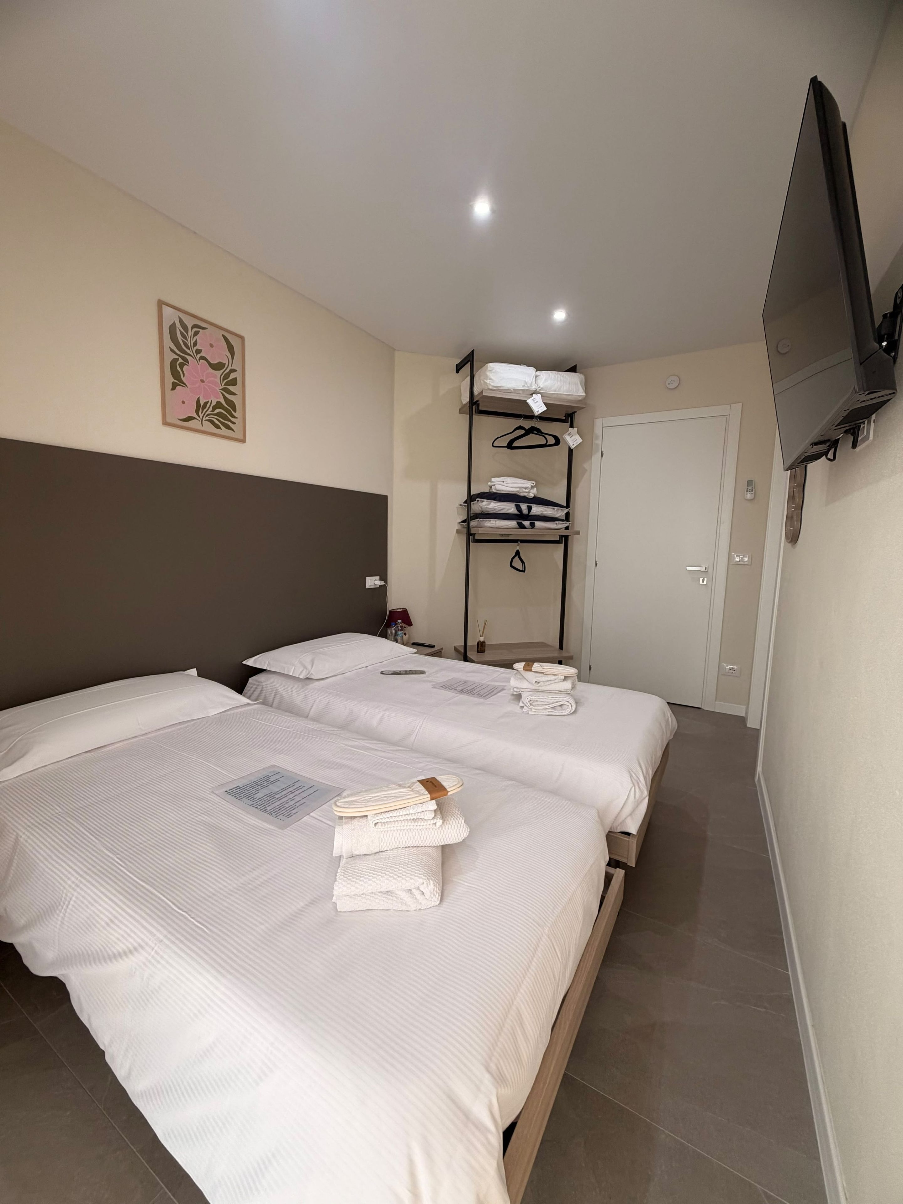 Ein Hotelzimmer mit zwei Betten, einem an der Wand montierten Fernseher, einem Nachttisch mit Lampe sowie einem Regal mit gefalteten Handtüchern und einer aufhängbaren Tasche.