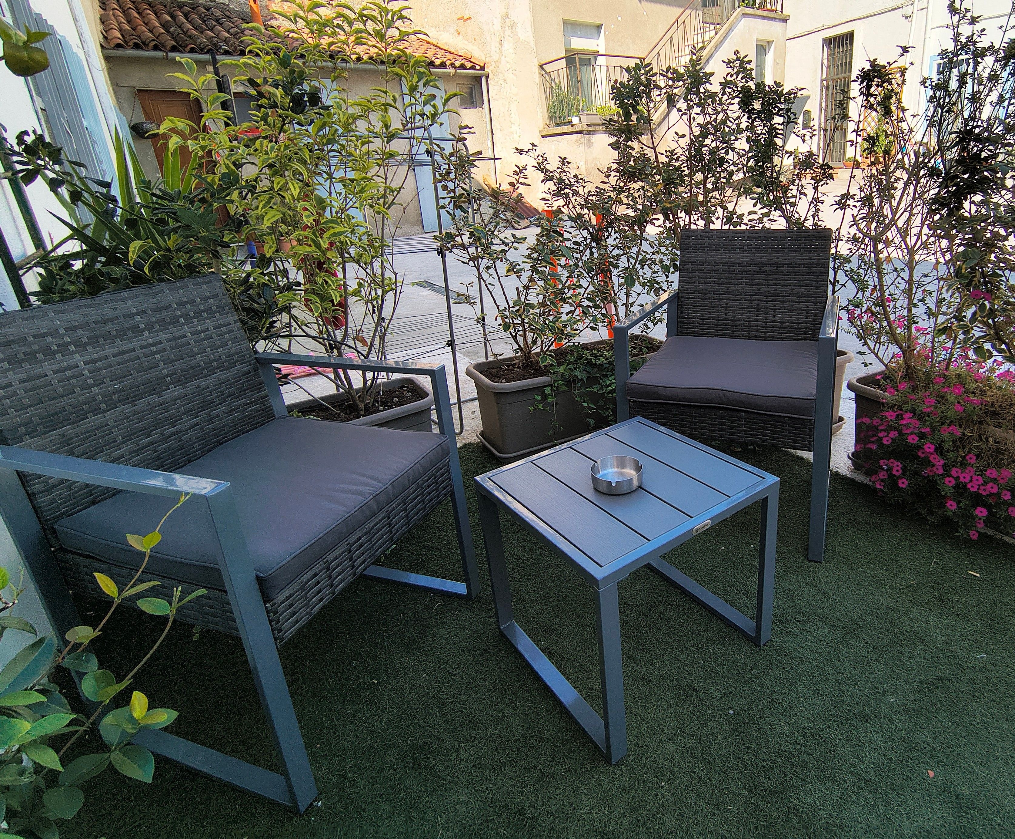 An outdoor seating area with two chairs and a small table, surrounded by potted plants and a view of a building.