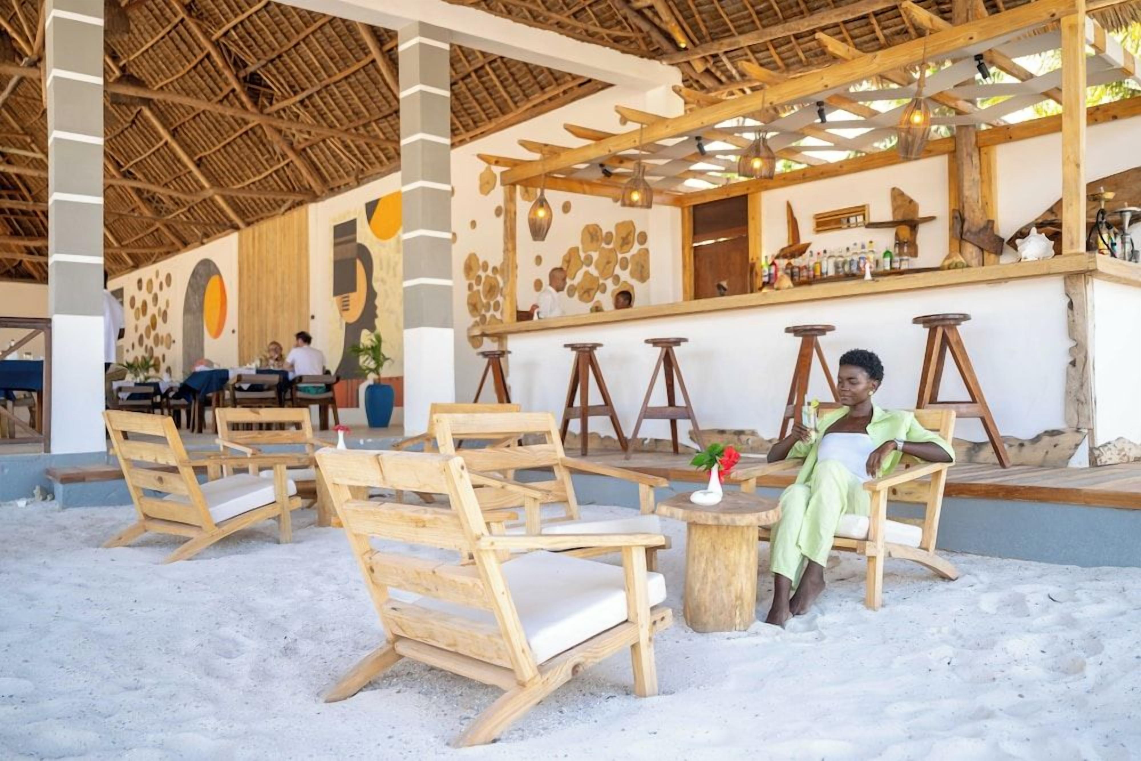 An outdoor seating area with wooden chairs and tables, a bar counter, and a thatched roof structure.