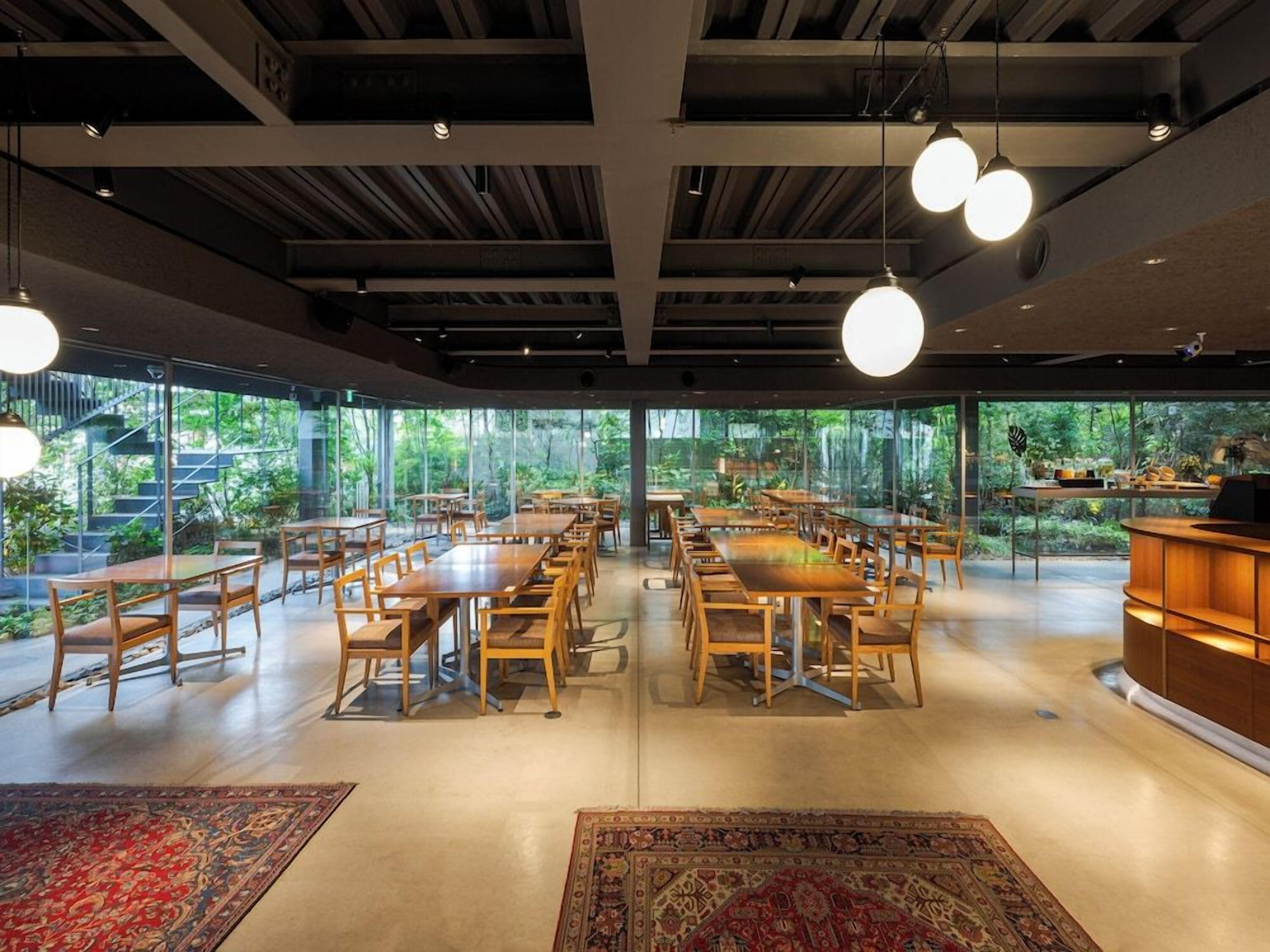 A spacious dining area with multiple tables and chairs, large windows offering a view of greenery, and hanging pendant lights.