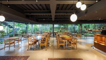 A spacious dining area with multiple tables and chairs, large windows offering a view of greenery, and hanging pendant lights.