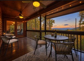 A wooden deck with wicker furniture, a table with a bottle and glasses, and a view of trees and mountains. - Big Mountain view cabin rental in Walnut Mountain. Pet Friendly, hot tub and pool table (Ellijay)