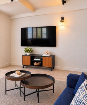 Ein modernes Wohnzimmer mit einem flachbildfernseher an der Wand, einem dunklen Holz-Konsolentisch mit Dekoration, zwei runden Couchtischen und einem blauen Sofa.