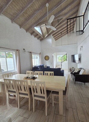 Une salle à manger spacieuse avec un plafond haut et un ventilateur de plafond, un salon avec un canapé et une télévision, et une vue sur l’extérieur par une fenêtre.