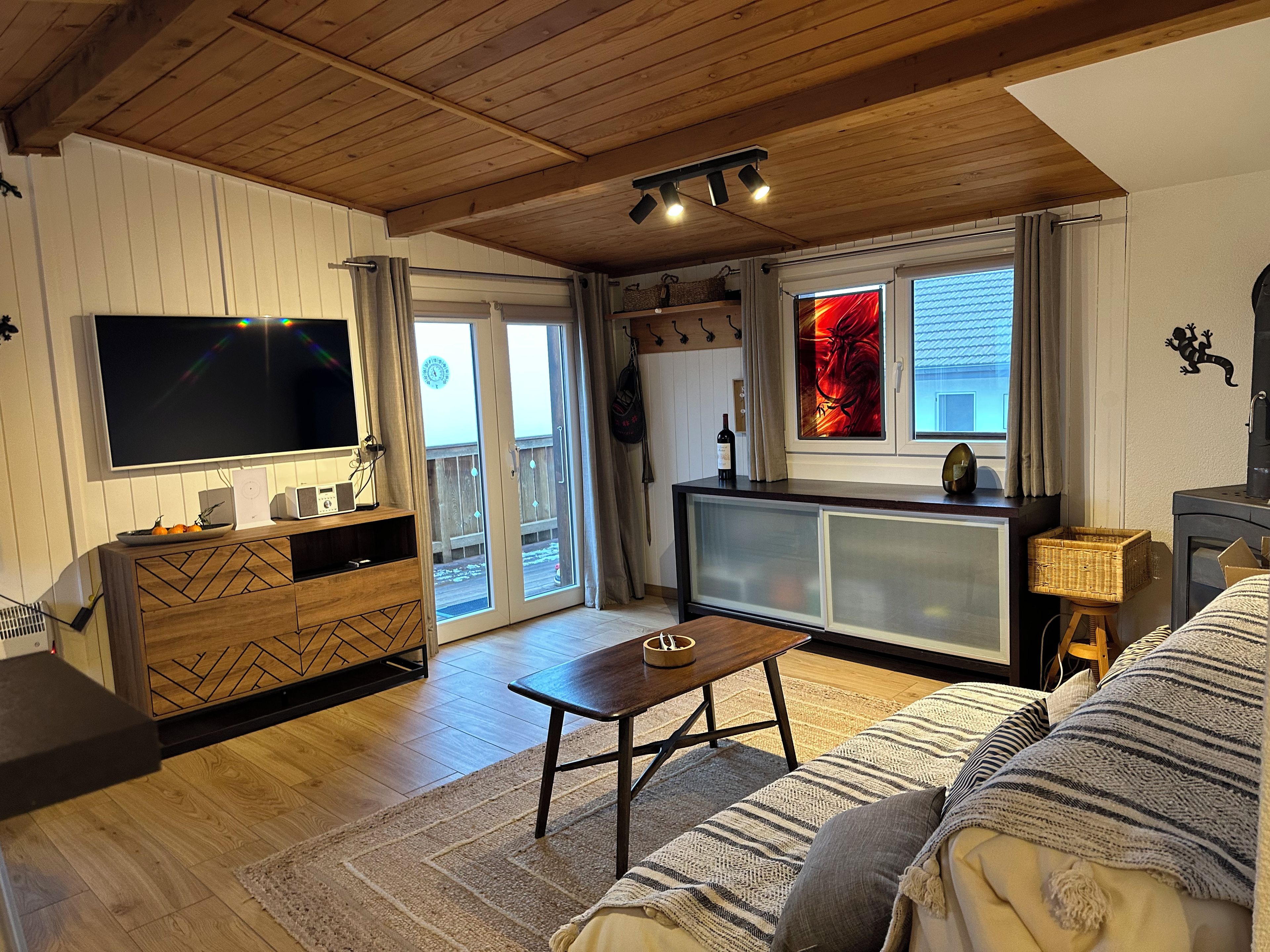 A cozy living room with a flat-screen TV, wooden furniture, a fireplace, and a balcony with a view.