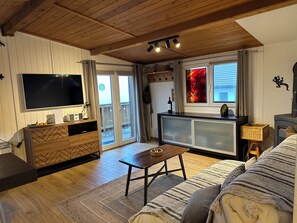 A cozy living room with a flat-screen TV, wooden furniture, a fireplace, and a balcony with a view.