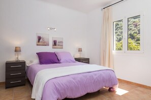 Un dormitorio con una cama, dos mesitas de noche, un ventanal con cortinas y dos lámparas de pared.