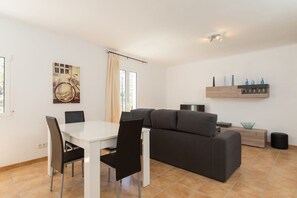 Una sala de estar moderna con una mesa de comedor blanca, sillas negras, un sofá gris y un televisor pequeño.