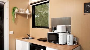 A compact kitchenette with a sink, microwave, and coffee maker, a window with a view of trees, and a shelf with glasses and a plant.