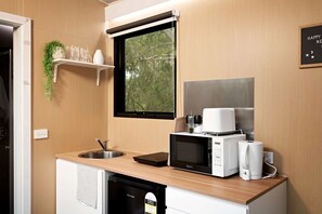 A compact kitchenette with a sink, microwave, and coffee maker, a window with a view of trees, and a shelf with glasses and a plant.
