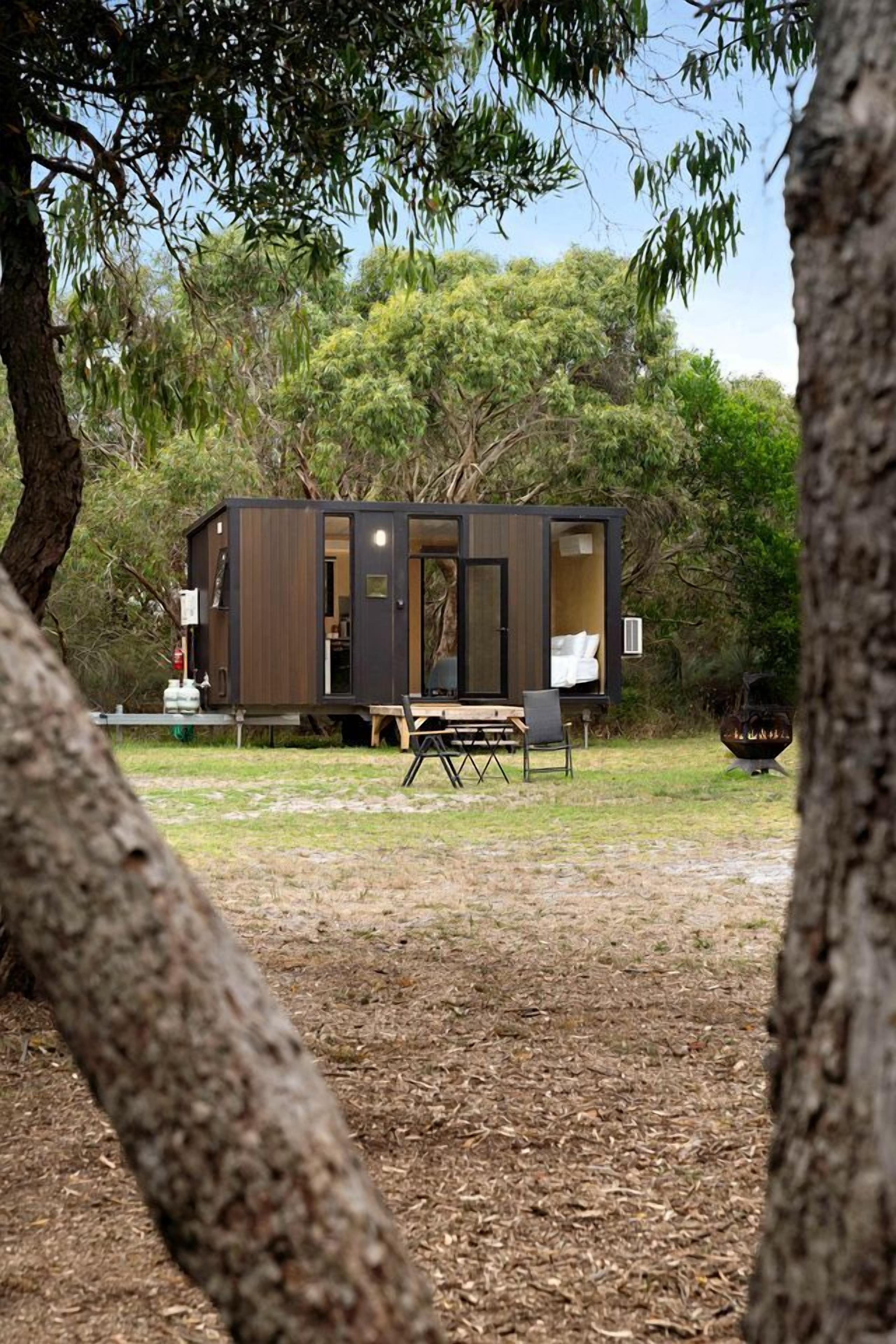 A small, modern cabin with an open door, a dining table, and a fire pit in a wooded area.