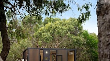 A small, modern cabin with an open door, a dining table, and a fire pit in a wooded area.