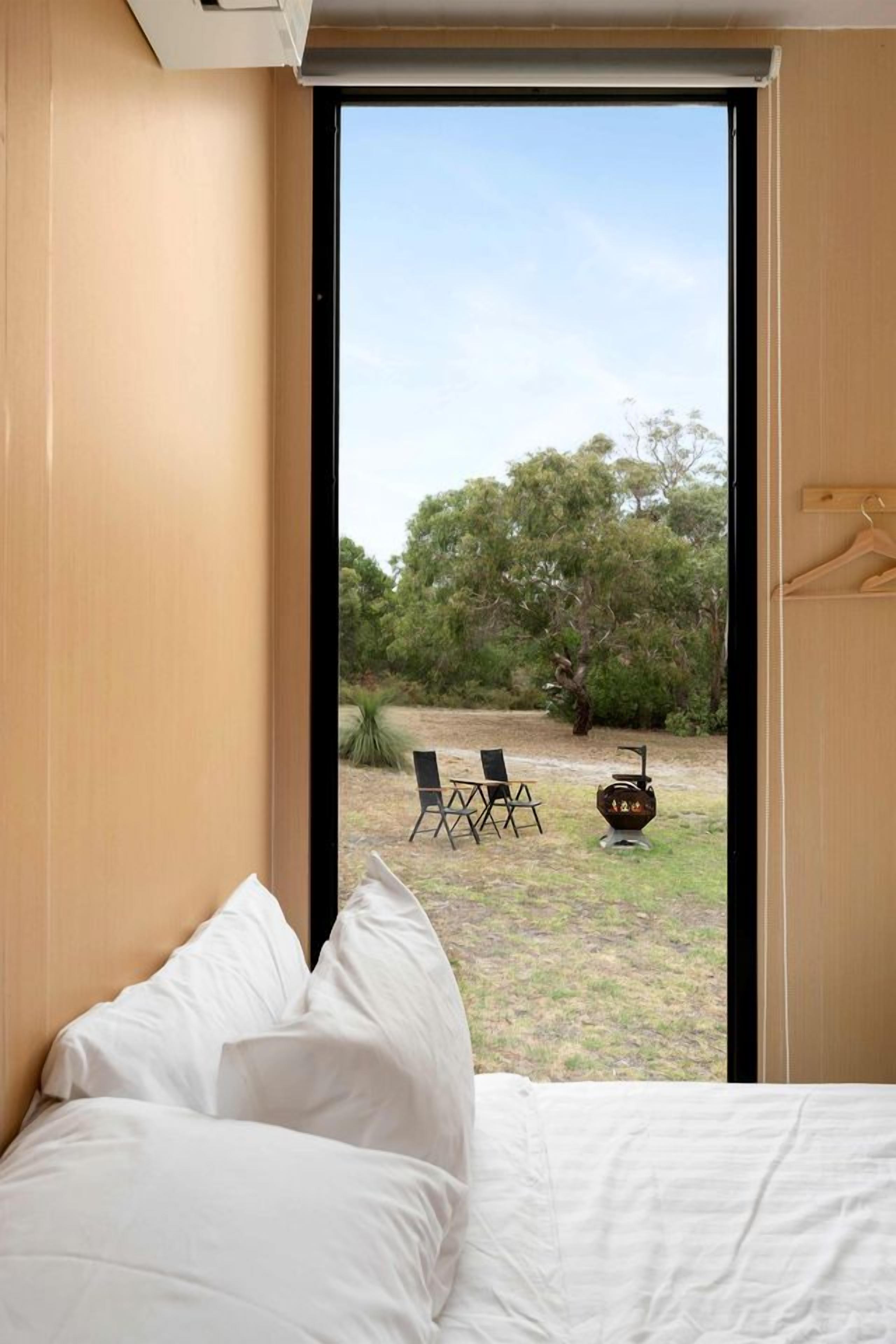 Ein Schlafzimmer mit Bett, Fenster und Blick auf einen Außenbereich mit Sitzgelegenheiten und einem Feuerkorb.