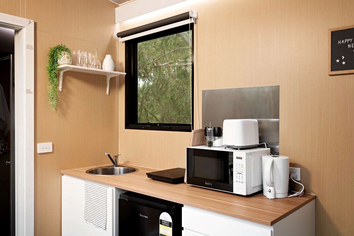 A compact kitchenette with a sink, microwave, and coffee maker, a window with a view of trees, and a shelf with glasses and a plant.