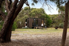 Une petite cabane de couleur sombre, dotée d’une porte coulissante, installée sur une sorte de remorque, dans un environnement naturel avec des arbres et une zone herbeuse.