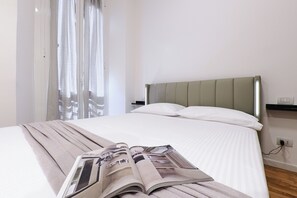 A neatly made bed with a magazine on it, a window with curtains, and a bedside table with a lamp. - Contempora - Cozy, Designer Two-Room Apartment for 2 | Monumentale & Sarpi M5 (Milano)