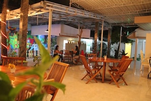 An indoor dining area with wooden tables and chairs, a high ceiling with metal beams, and a colorful mural on the wall. - Hotel Wisata Baru (Serang)