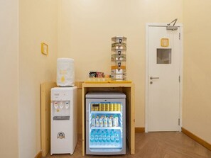 A compact kitchenette with a water dispenser, a mini-fridge stocked with beverages, and a small shelf with snacks. - Bloom Hotel - Saki Vihar Powai (Mumbai)