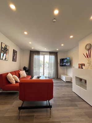 Un salon moderne avec un canapé rouge, une table basse, un téléviseur à écran plat fixé au mur et une grande fenêtre avec des rideaux.