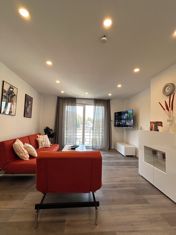 A modern living room with a red sofa, a coffee table, a flat-screen TV mounted on the wall, and a large window with curtains.