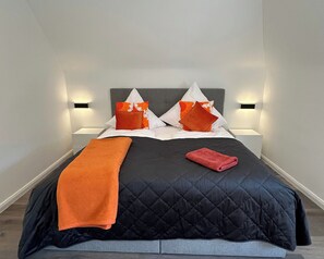 A bed with a dark quilted bedspread, two pillows with orange and white patterns, and a red towel.