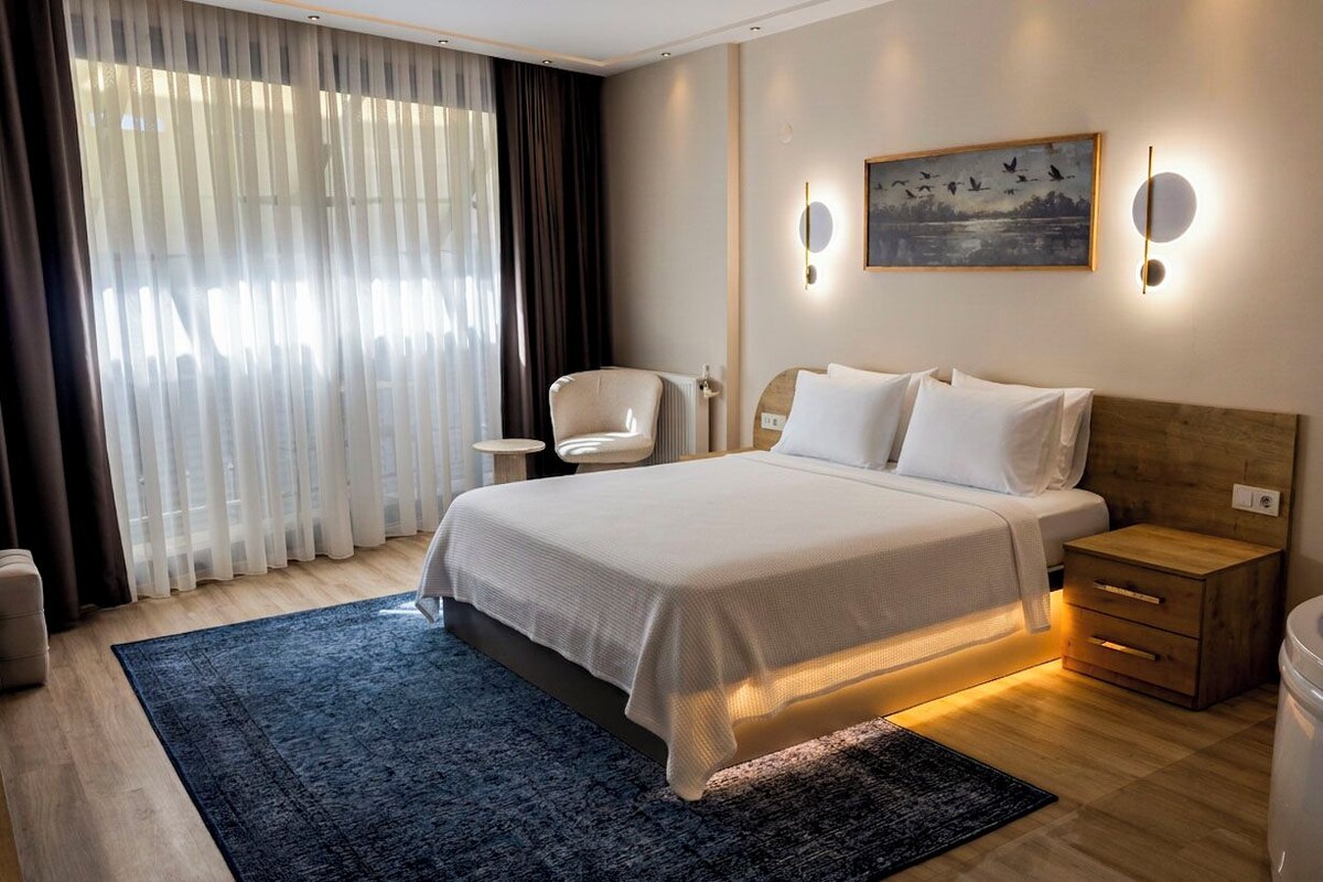 A hotel room with a large bed, a bedside table, a chair, a nightstand, and a window with curtains.