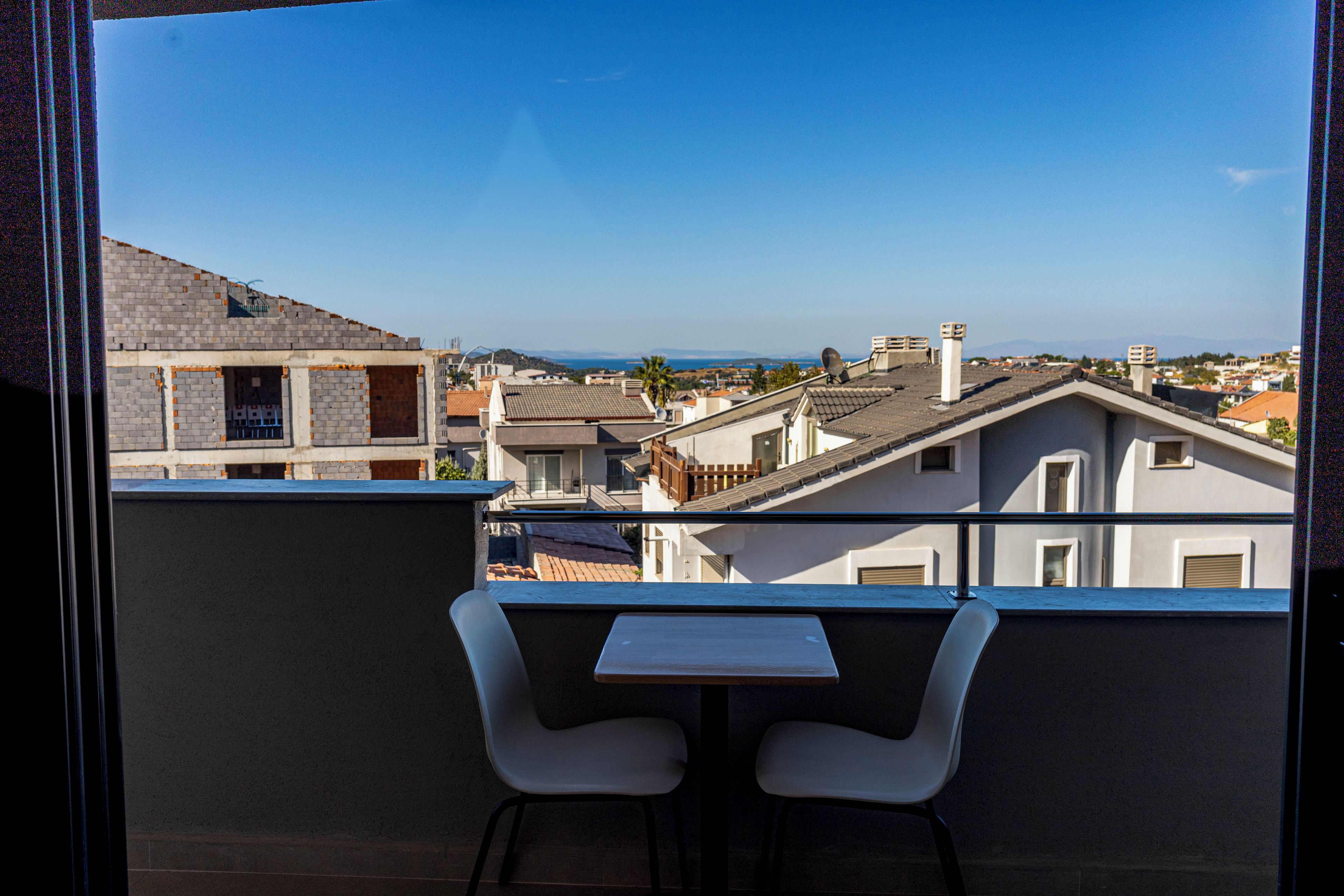 Un balcón con dos sillas blancas y una mesita, con vistas a una zona residencial con varios edificios y un cielo despejado.