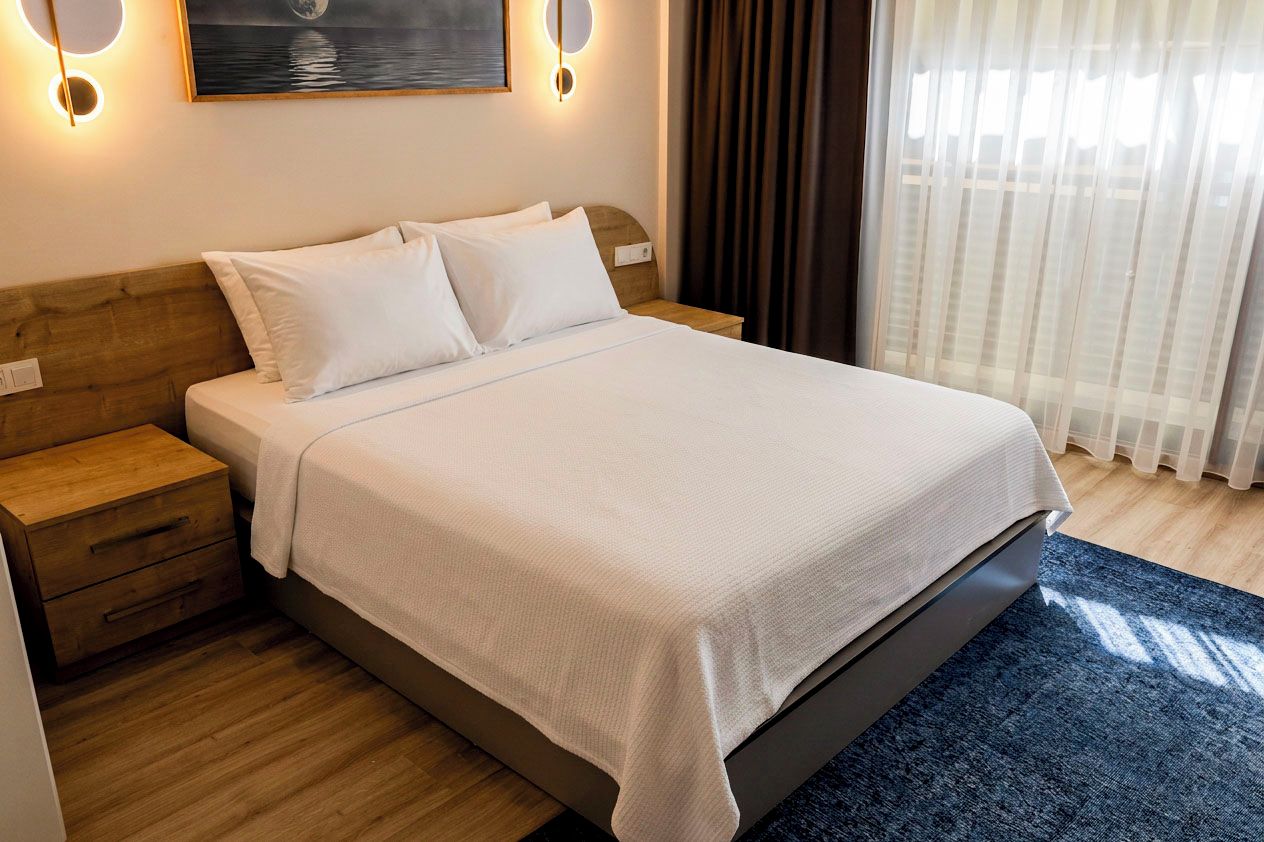 Una cama bien hecha con cubrecama y almohadas blancas, un cabecero de madera, una mesita de noche con cajones y una lámpara de pared.