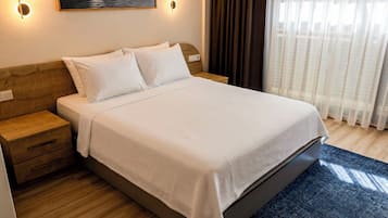 Una cama bien hecha con cubrecama y almohadas blancas, un cabecero de madera, una mesita de noche con cajones y una lámpara de pared.