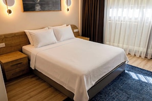 Una cama bien hecha con cubrecama y almohadas blancas, un cabecero de madera, una mesita de noche con cajones y una lámpara de pared.