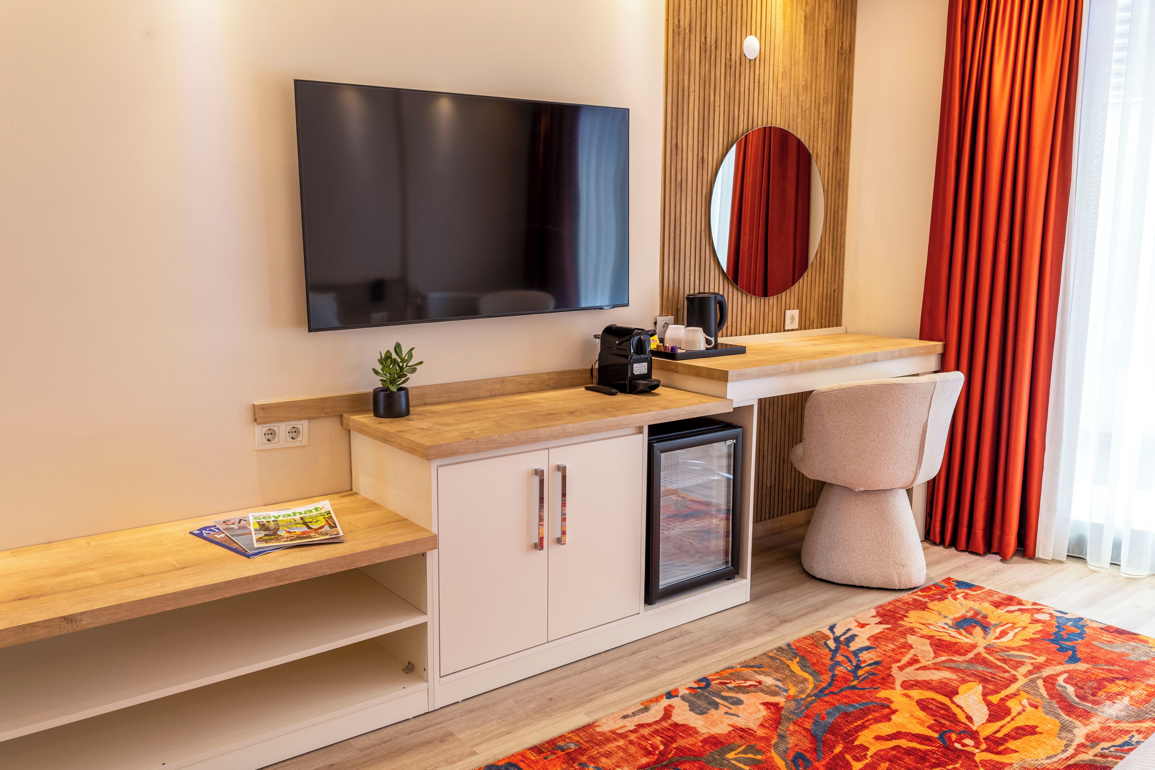 Una habitación de hotel moderna con una televisión de pantalla plana, un refrigerador pequeño, una cafetera, una mesita de noche con una revista y un gran ventanal con cortinas.
