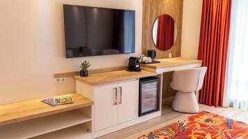 Una habitación de hotel moderna con una televisión de pantalla plana, un refrigerador pequeño, una cafetera, una mesita de noche con una revista y un gran ventanal con cortinas.