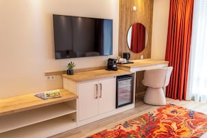 Una habitación de hotel moderna con una televisión de pantalla plana, un refrigerador pequeño, una cafetera, una mesita de noche con una revista y un gran ventanal con cortinas.