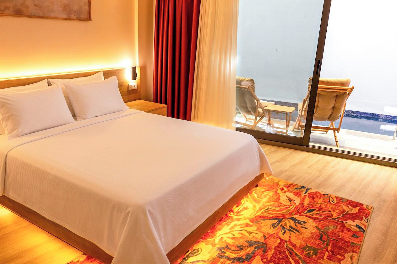Habitación de hotel con una cama grande, piso de madera, alfombra con estampado rojo y blanco, una mesita de noche, una lámpara, una silla y vistas al mar a través de una puerta de cristal.