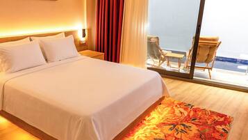 Habitación de hotel con una cama grande, piso de madera, alfombra con estampado rojo y blanco, una mesita de noche, una lámpara, una silla y vistas al mar a través de una puerta de cristal.