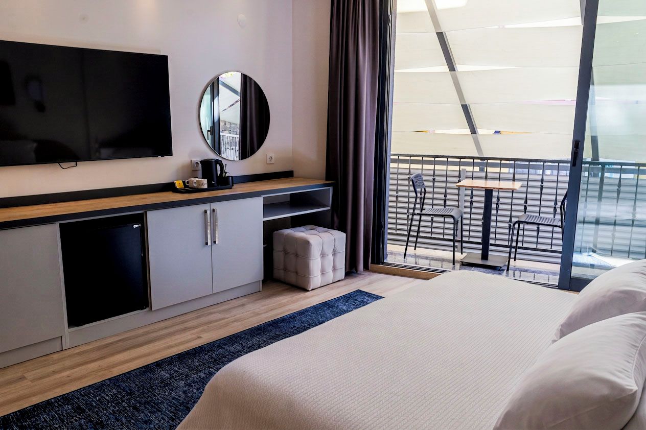 A hotel room with a bed, a TV, a mirror, a small table with a chair, and a balcony with a railing.
