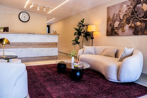 Un área de recepción moderna con un mostrador de mármol, un reloj, un cartel de recepción, un sofá, una mesa de centro y una planta grande.