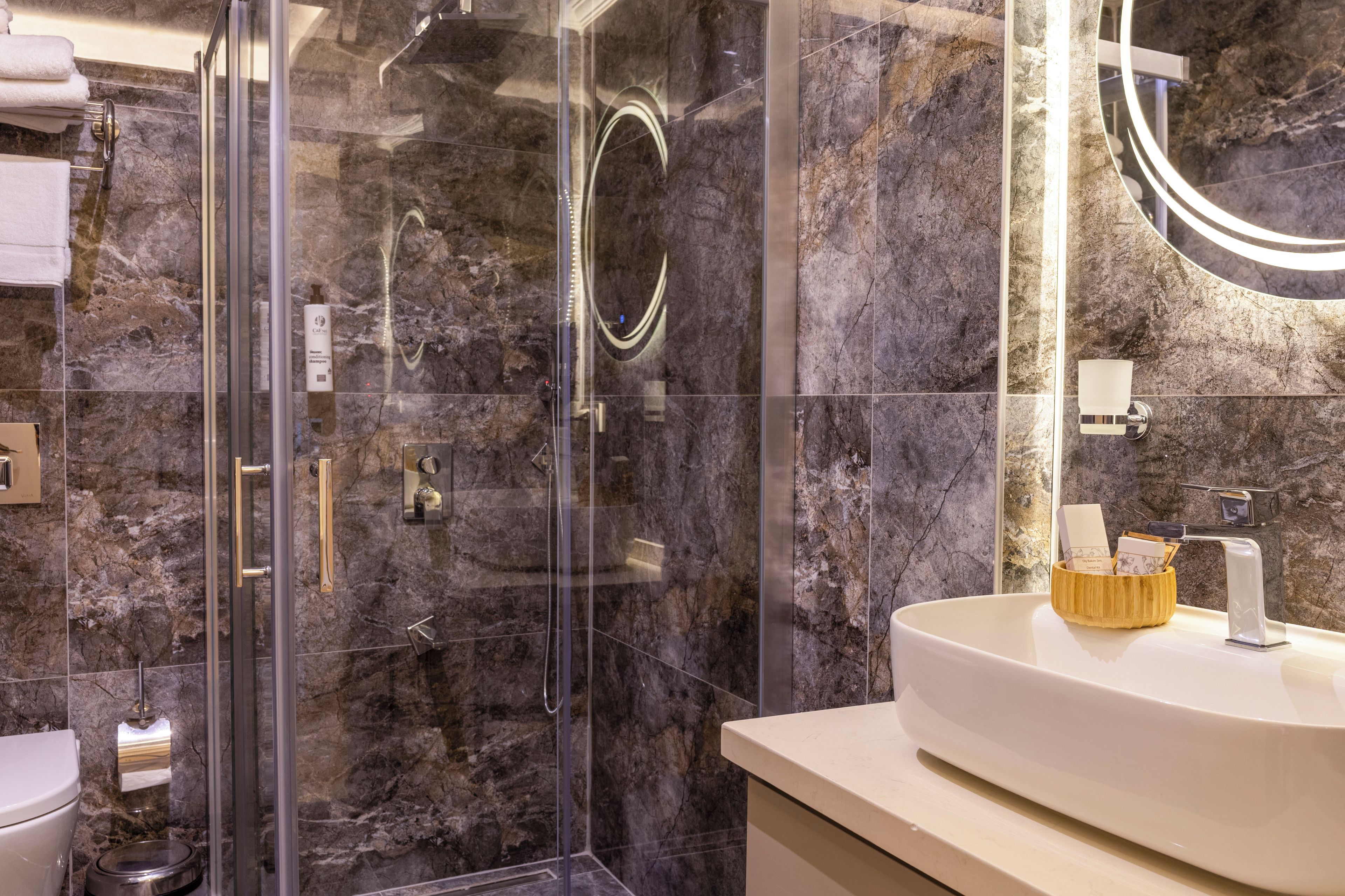 Un baño moderno con ducha de vidrio, paredes de mármol y lavamanos blanco con un cuenco de madera.