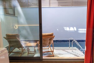 A room with a view of a pool, two chairs, a small table, and a metal ladder.
